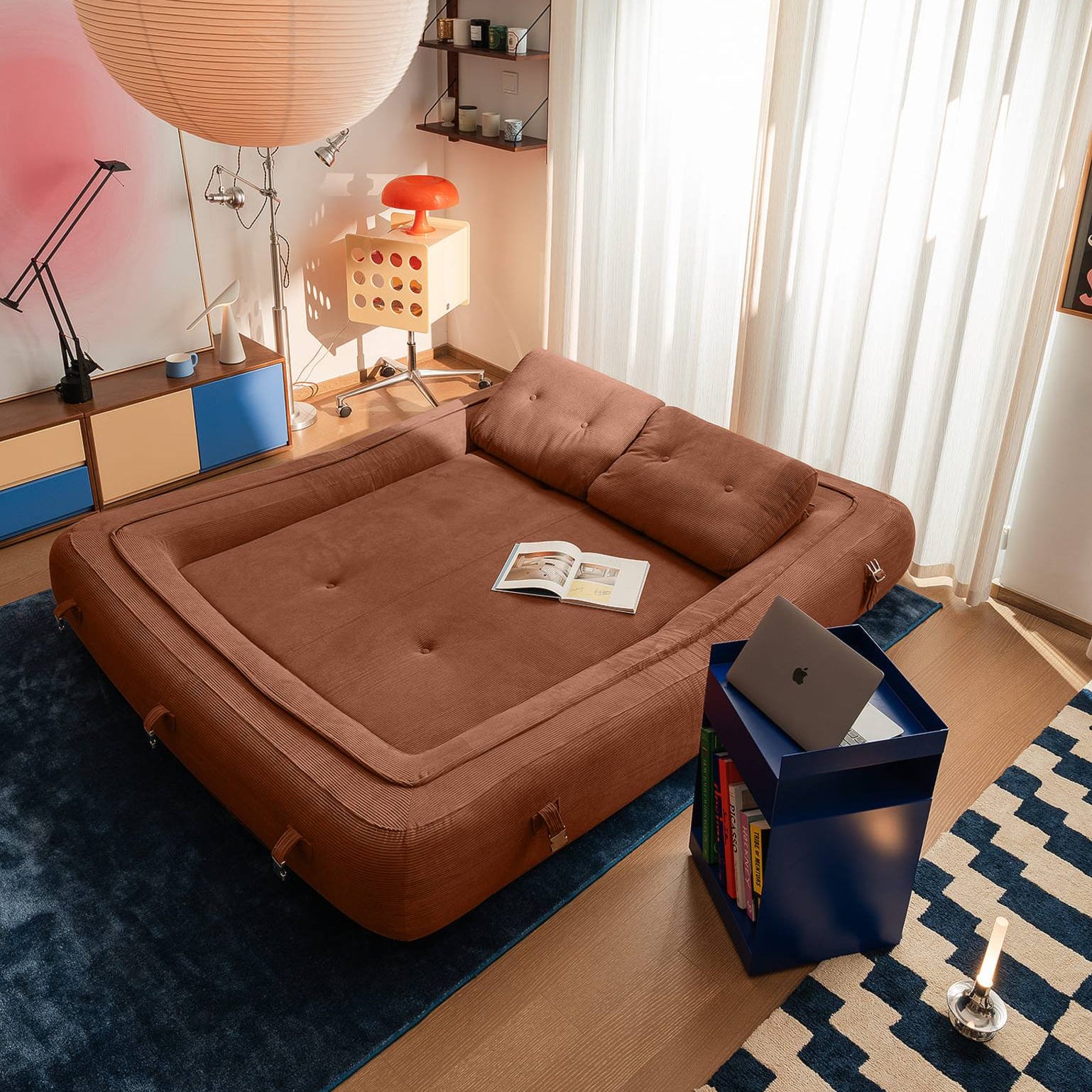 Brown sofa bed in a living room with a lamp and books on a blue rug.