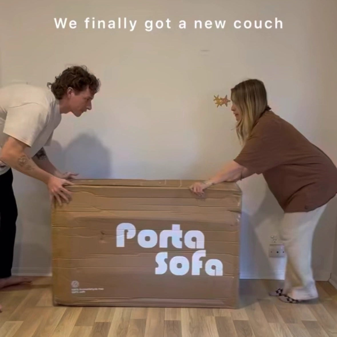 Two people lifting a 'Porta Sofa' box in a room.
