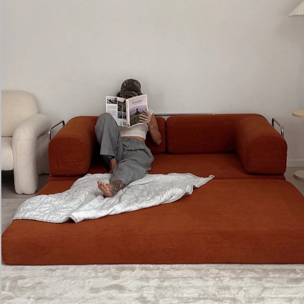 Person lying on a large orange sofa reading a magazine in a minimalistic room.
