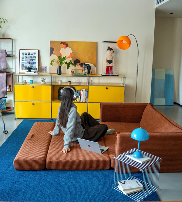 Living room with a child on a brown sofa using a laptop, yellow cabinet, and colorful decor.
