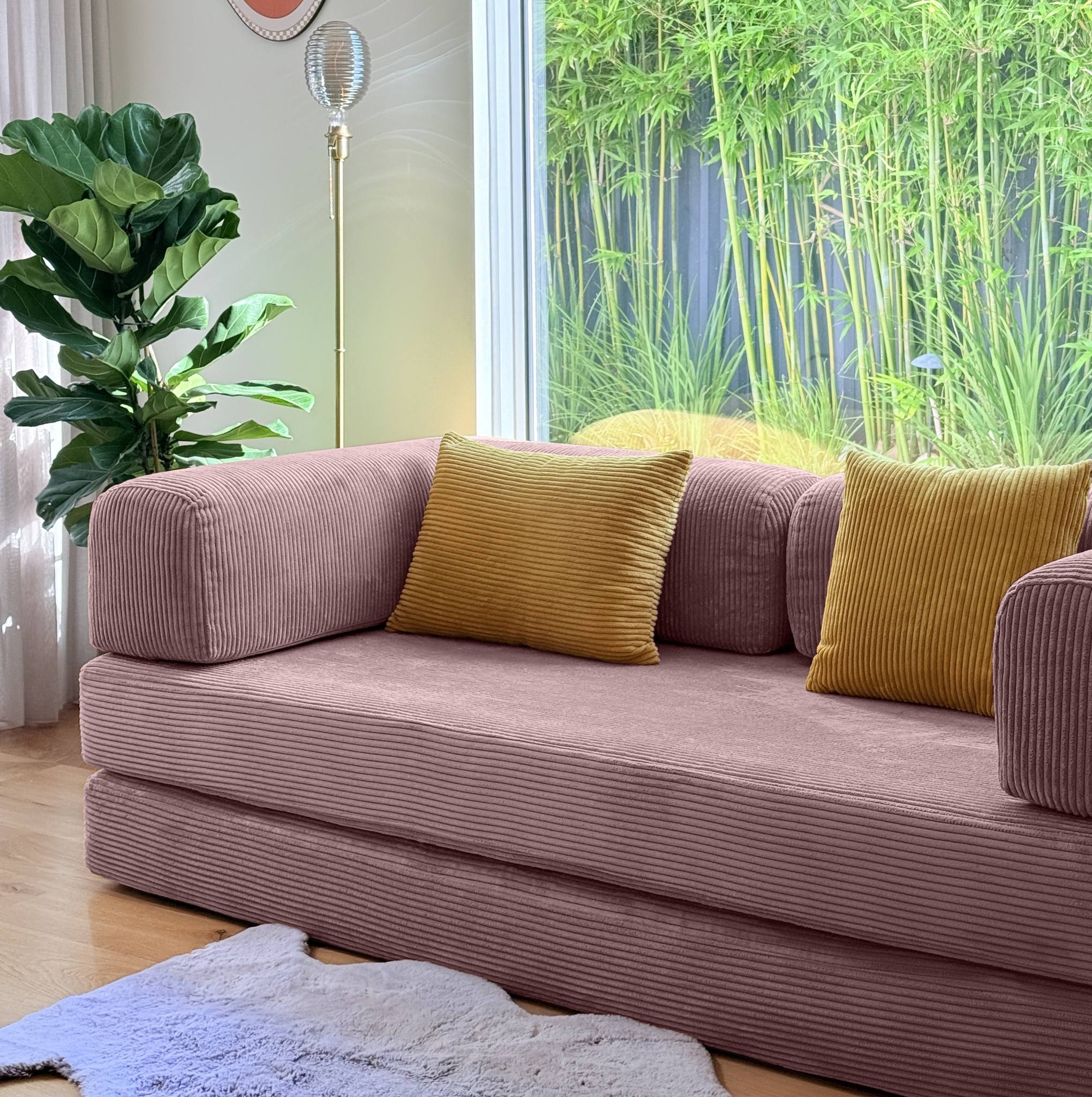 Pink sofa with yellow cushions in a room with bamboo plants outside.