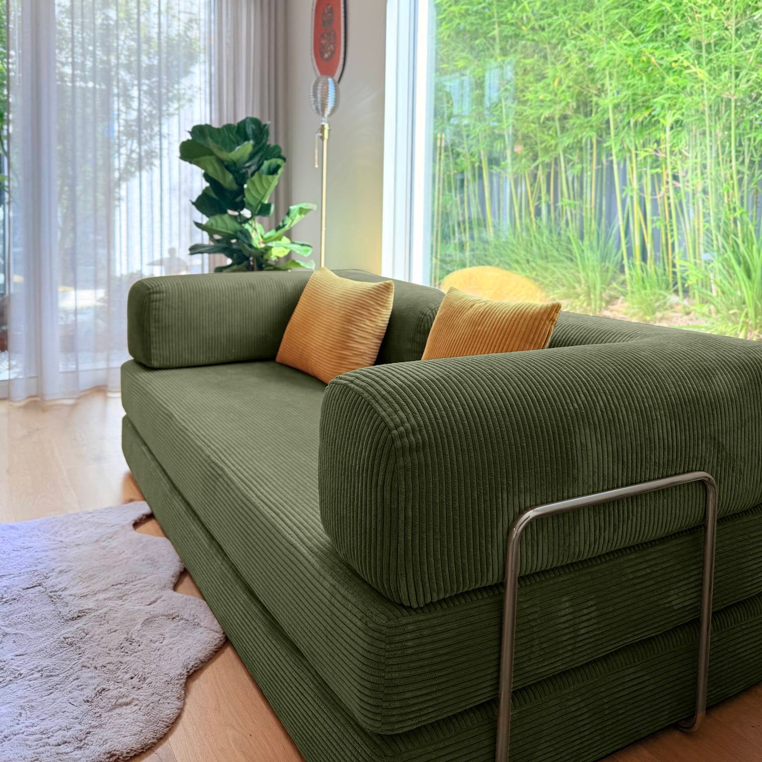 Green sofa with two yellow cushions in a room with large windows showing greenery outside.