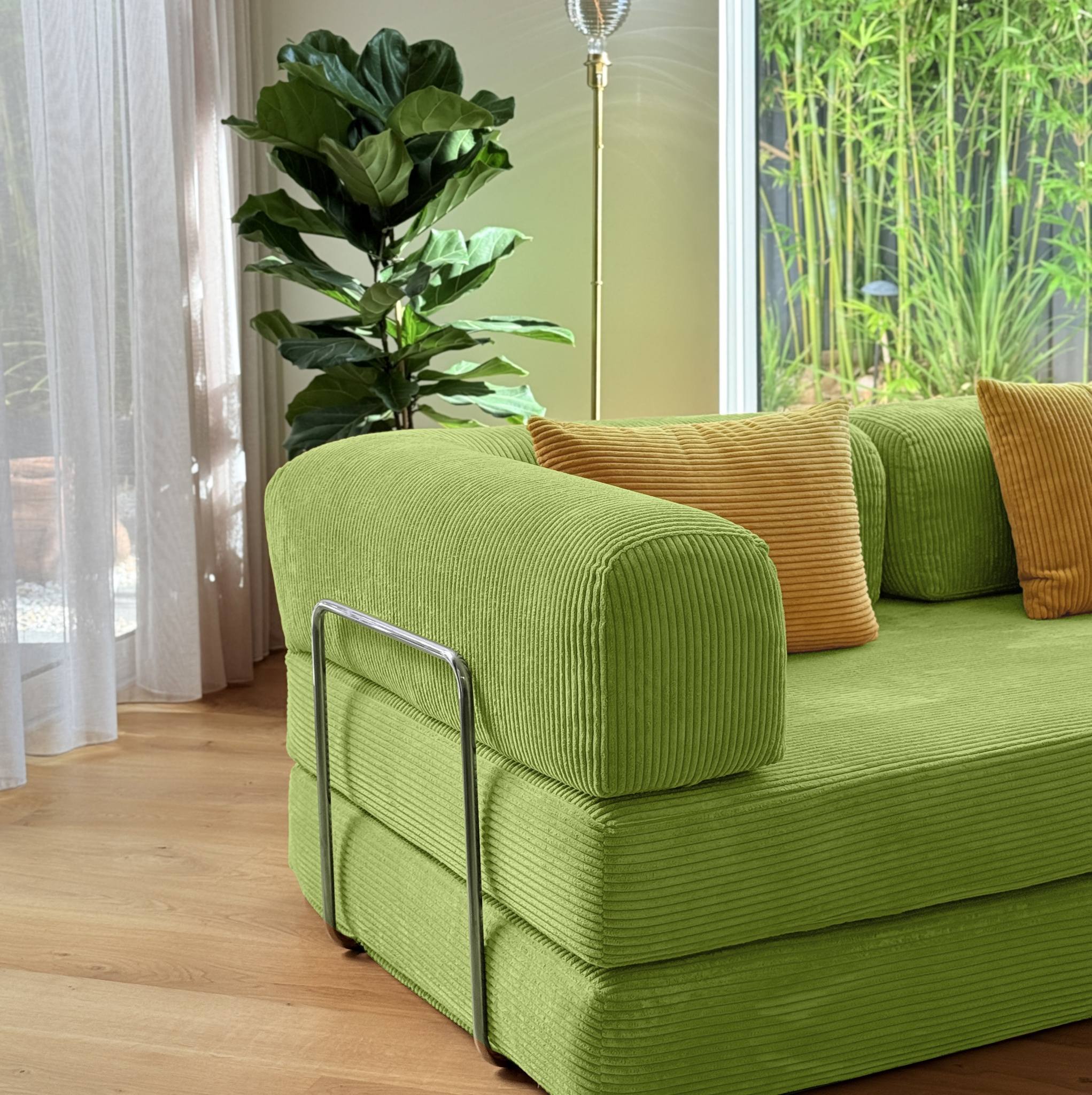 Green sofa with orange cushions in a room with plants and curtains