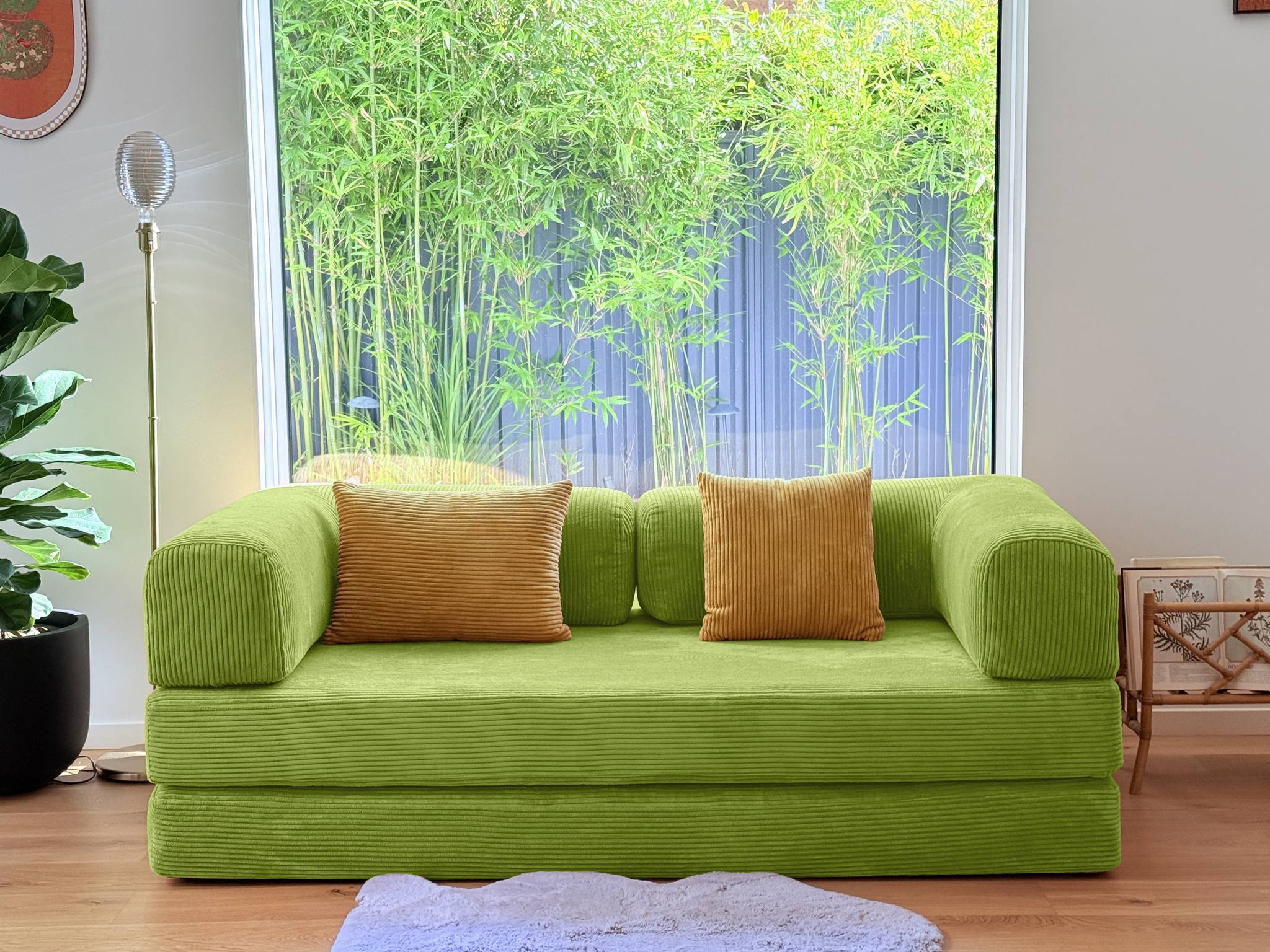 Green sofa with orange cushions in a room with a large window and plants.