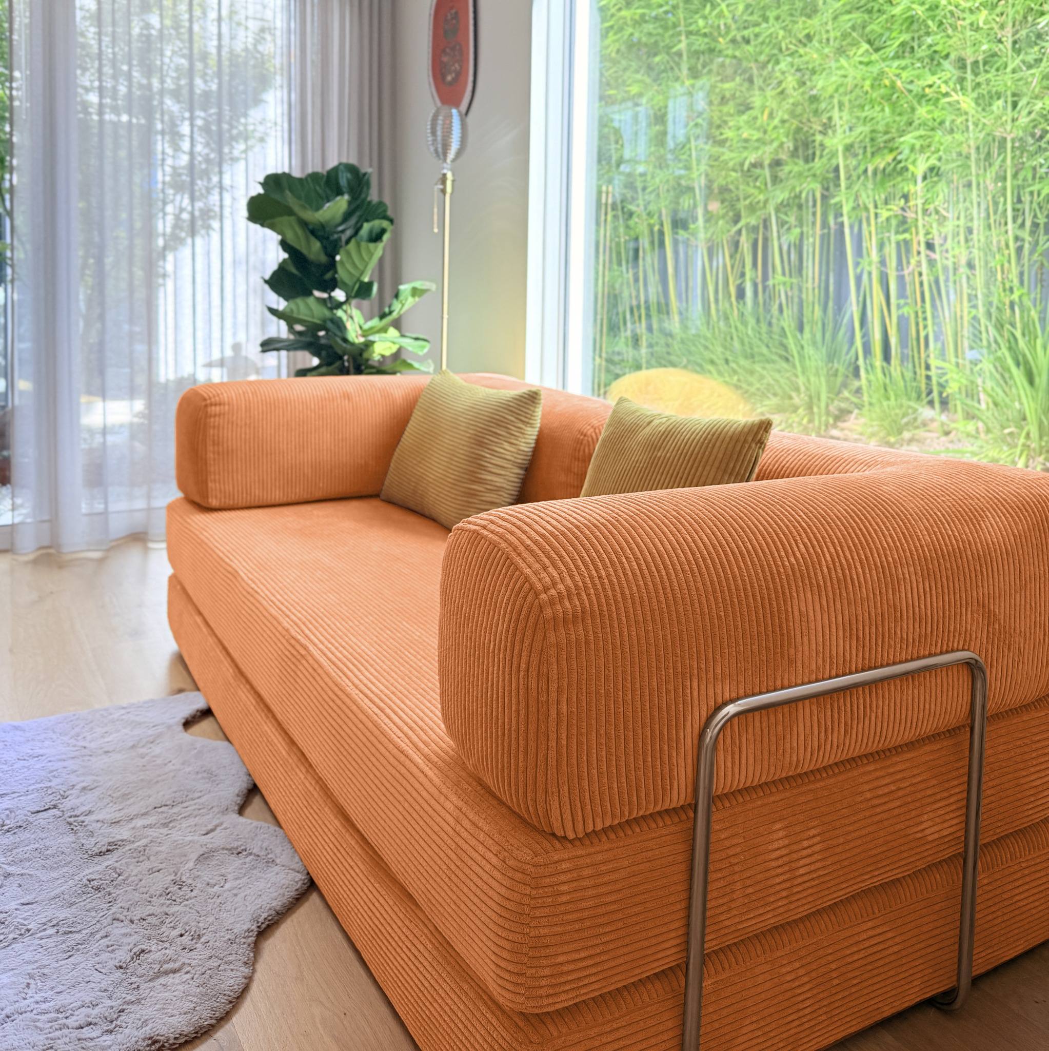 Orange sofa with two green pillows in a room with large windows and plants.