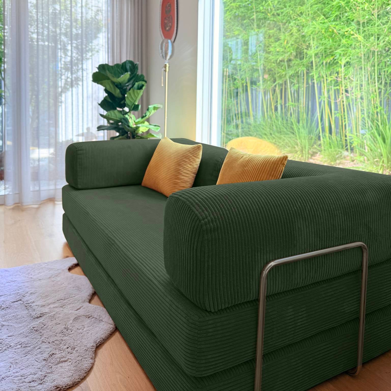 Green sofa with gold cushions in a room with large windows showing greenery outside.