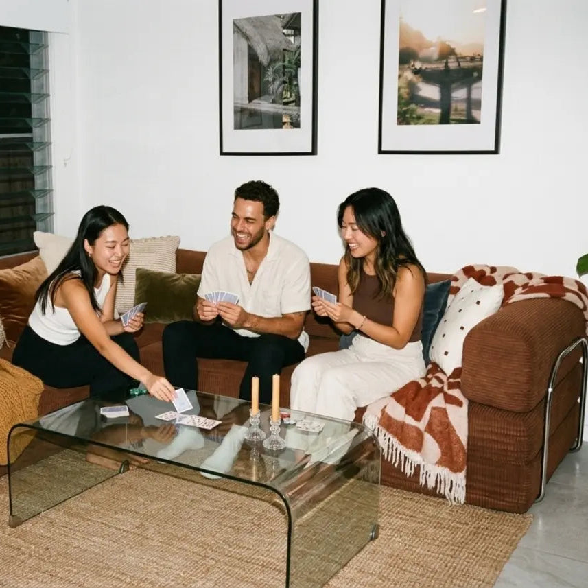 Three people sitting on a brown couch in a living room with a glass coffee table.
